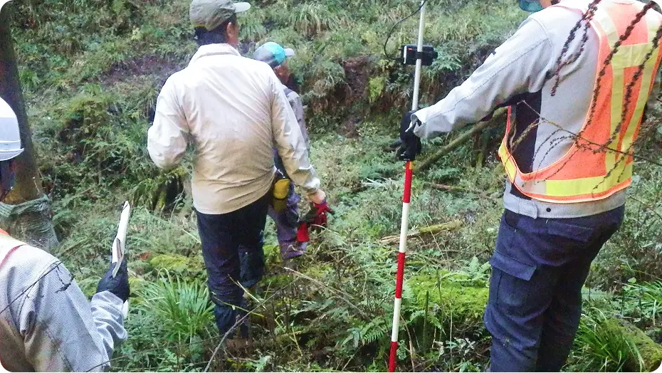 現地調査の様子：ヘルメットを着用した調査員たちが、測量ポールや機材を持って山林に入り、地形や位置の測量調査を行っている風景